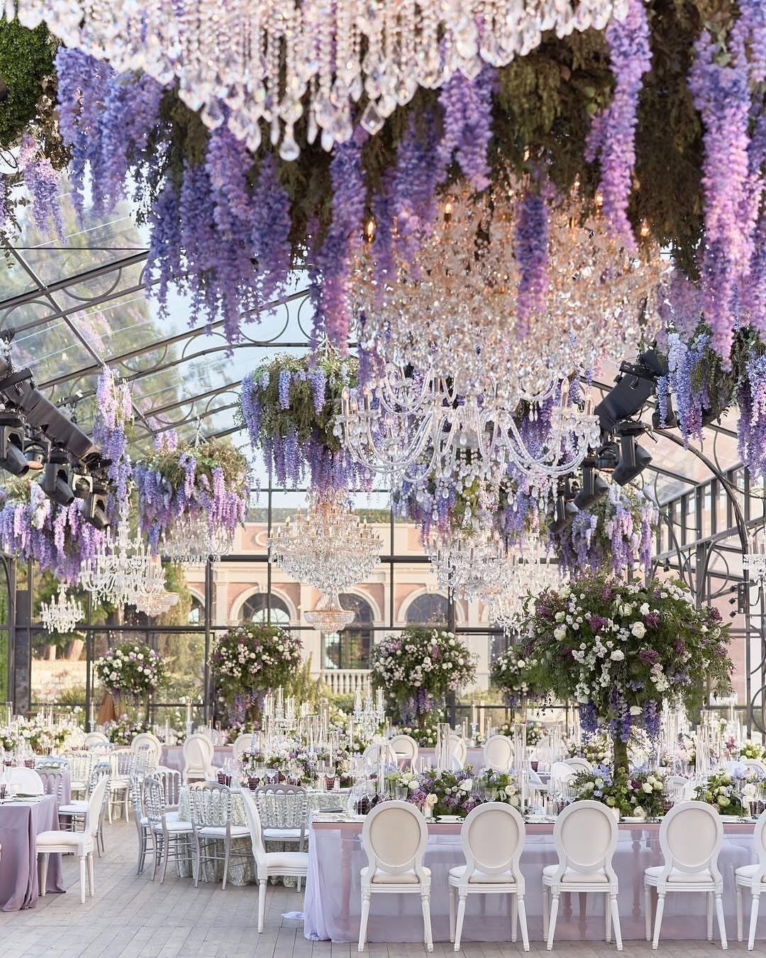 Lavender wedding reception with hanging wisteria, crystal chandeliers, and white chairs.