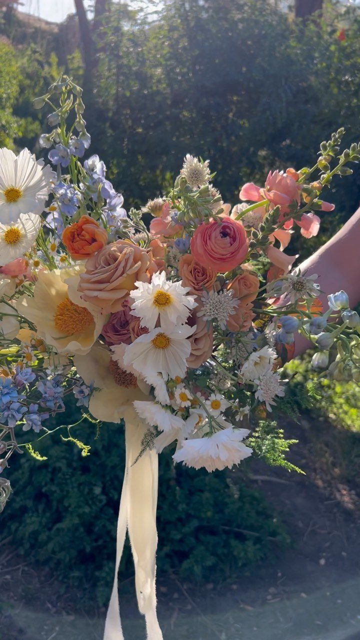 Bridal bouquet with peach roses, white daisies, and lavender blue wildflowers tied with ribbon.