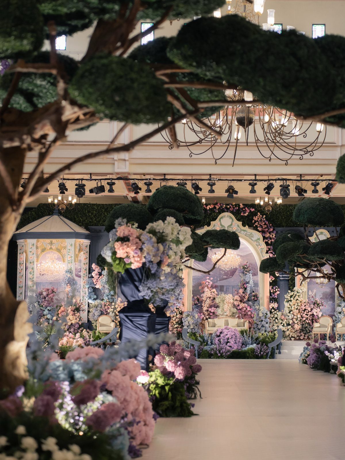 Lavish wedding reception venue with purple flowers, green topiaries, and a crystal chandelier.