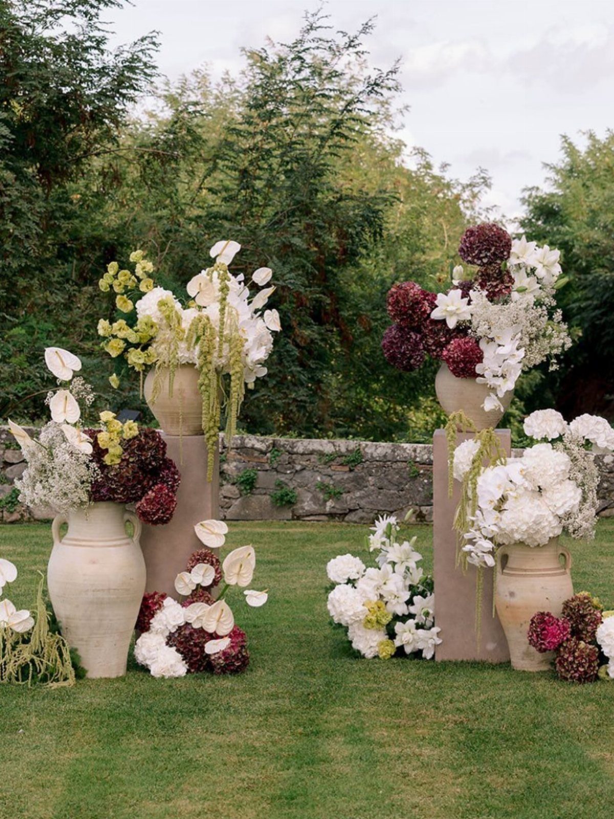 Terracotta vases with white and burgundy flowers on pedestals in a lush green garden.
