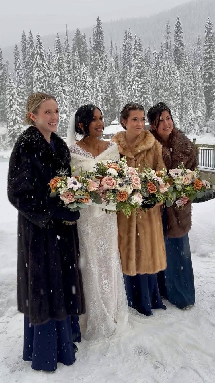 Four women in formal attire and fur coats posing in a snowy pine forest.