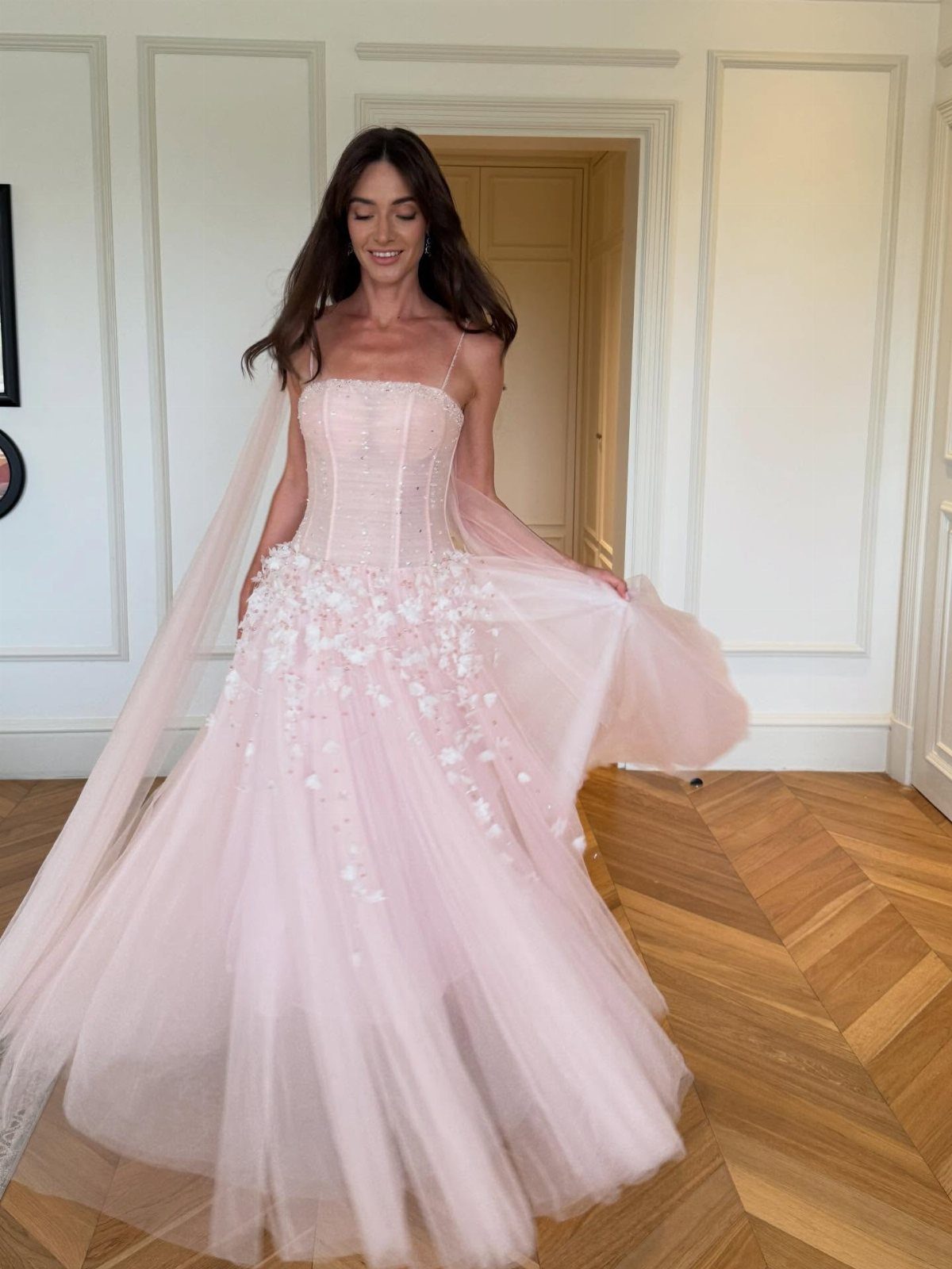 A woman in a pink floral tulle gown smiling in a white paneled room.