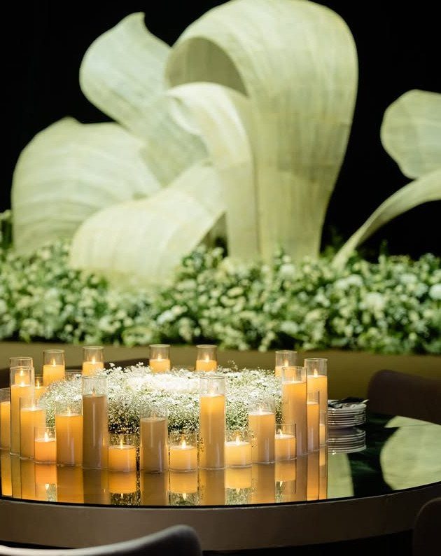 25 Wedding Stuff Ideas for Your 2026 Ceremony and Party 22 Glowing cylindrical glass candles surrounding a white baby's breath floral centerpiece on a reflective table.