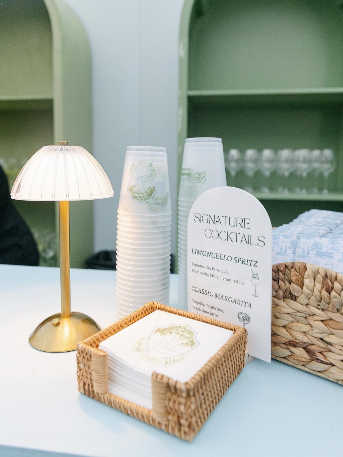 25 Wedding Stuff Ideas for Your 2026 Ceremony and Party 9 Signature cocktail menu sign next to stacked paper cups and a small gold lamp on a table.