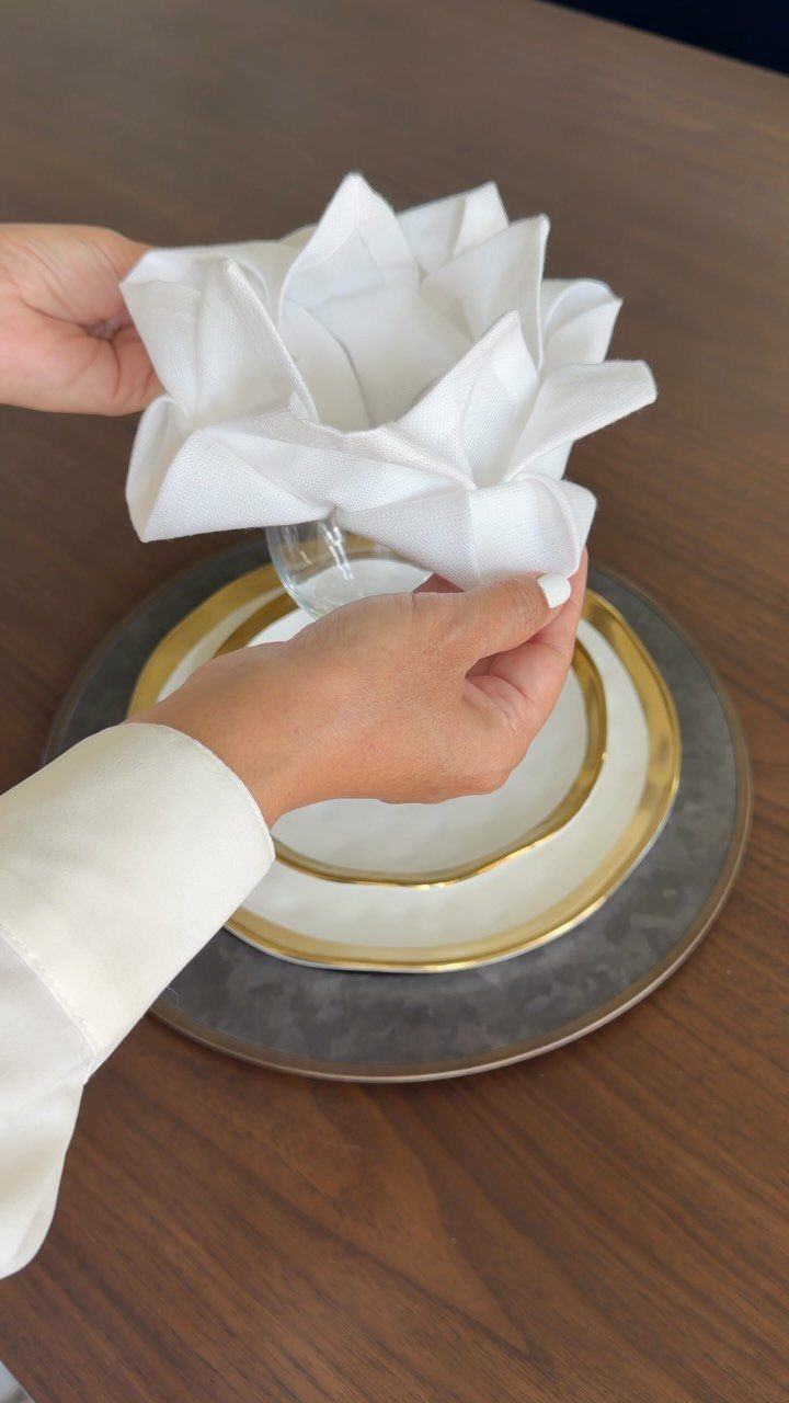 25 Wedding Stuff Ideas for Your 2026 Ceremony and Party 18 Hands placing a white cloth napkin folded into a flower shape onto a gold-rimmed plate.