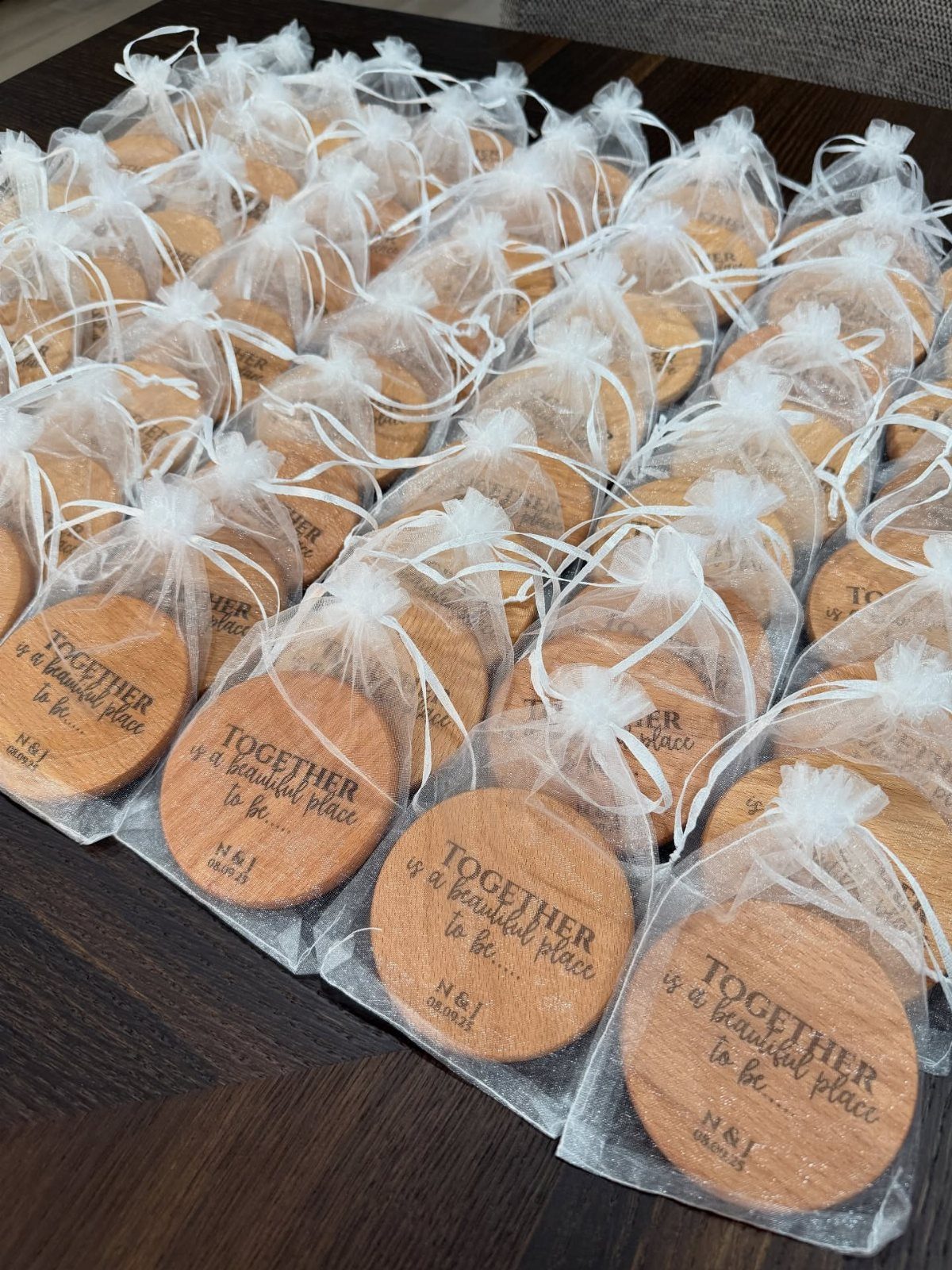 25 Wedding Stuff Ideas for Your 2026 Ceremony and Party 23 Rows of wooden coasters engraved with text, wrapped in white organza drawstring bags on a dark table.