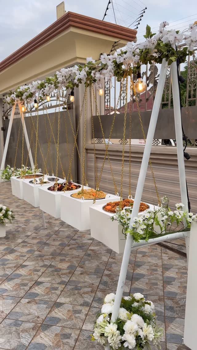 25 Wedding Stuff Ideas for Your 2026 Ceremony and Party 17 White pedestals displaying various food platters under a floral decorated frame with hanging lights.