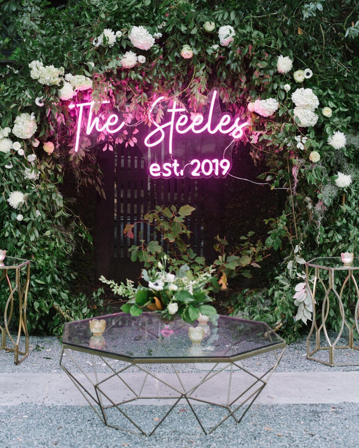 25 Wedding Stuff Ideas for Your 2026 Ceremony and Party 6 Pink neon sign reading The Steeles mounted on a lush green floral hedge backdrop.