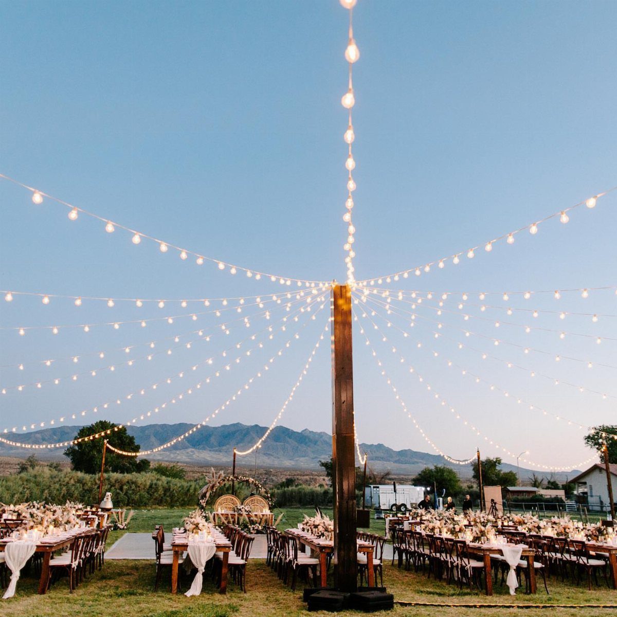 25 Wedding Stuff Ideas for Your 2026 Ceremony and Party 4 Long dining tables set outdoors under many radiating strands of warm string lights at twilight.