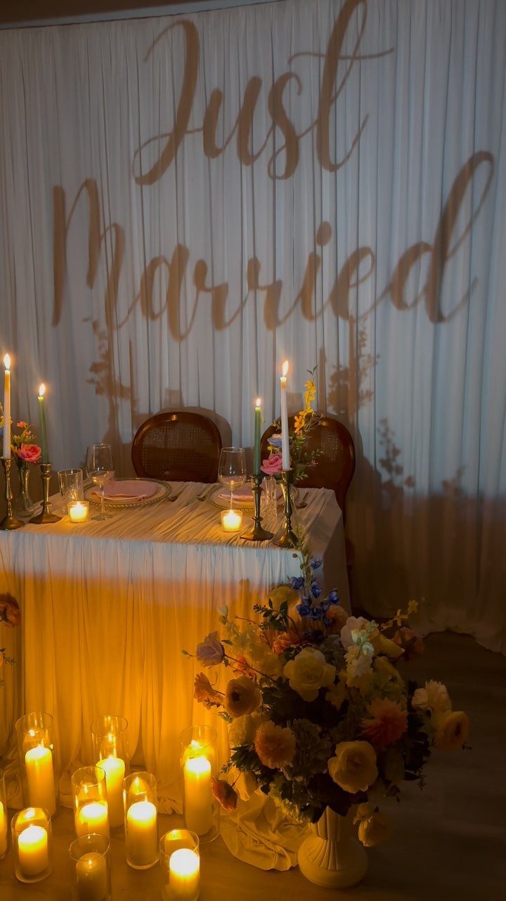 25 Wedding Stuff Ideas for Your 2026 Ceremony and Party 14 A sweetheart table decorated with white linens, candles, and flowers against a Just Married shadow backdrop.