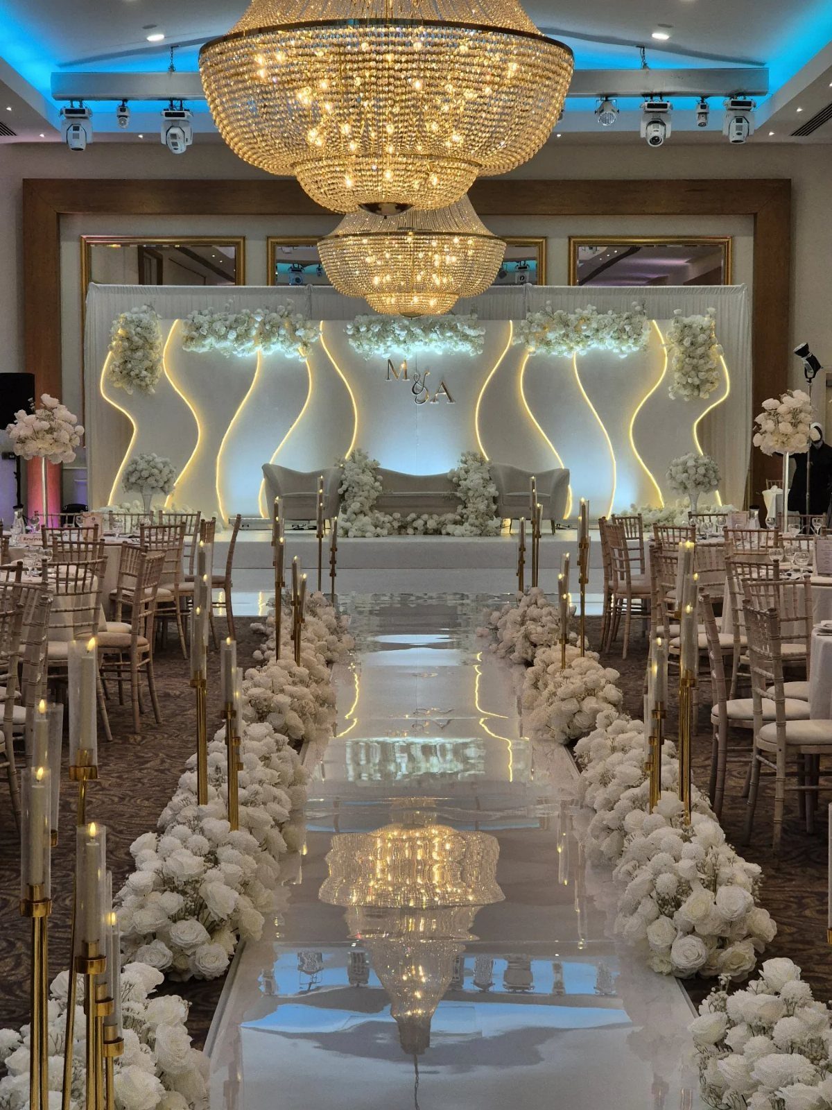 Wedding Theme and Motif: 16 Fresh Ideas for 2026 15 A white wedding aisle with reflective flooring, white flowers, and gold candle holders under a chandelier.