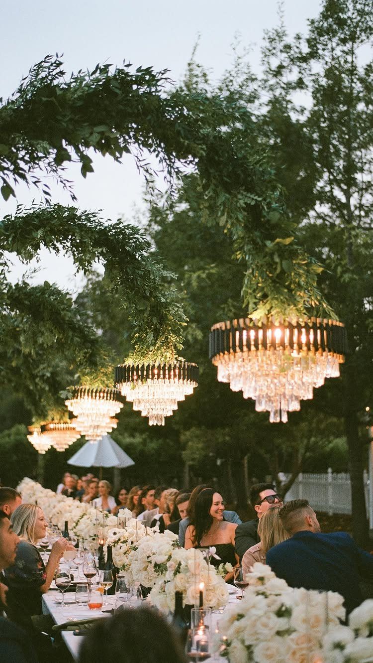 Wedding Themes 2026: 12 Ideas You Will Actually Want to Steal 13 Guests dine at a long wedding table under leafy arches and glowing crystal chandeliers.