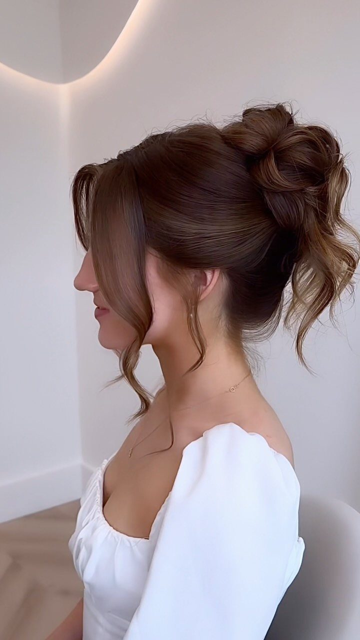 27 Bridesmaid Wedding Updo Hairstyles from Buns to Braids 4 Woman with brown hair in a voluminous high bun and curled face-framing pieces.