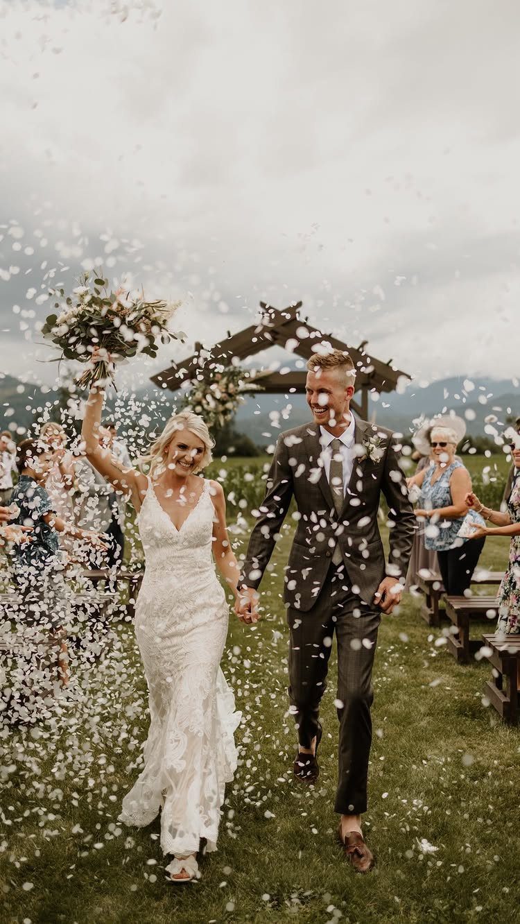 Bride and groom walking through a shower of white confetti petals outdoors.