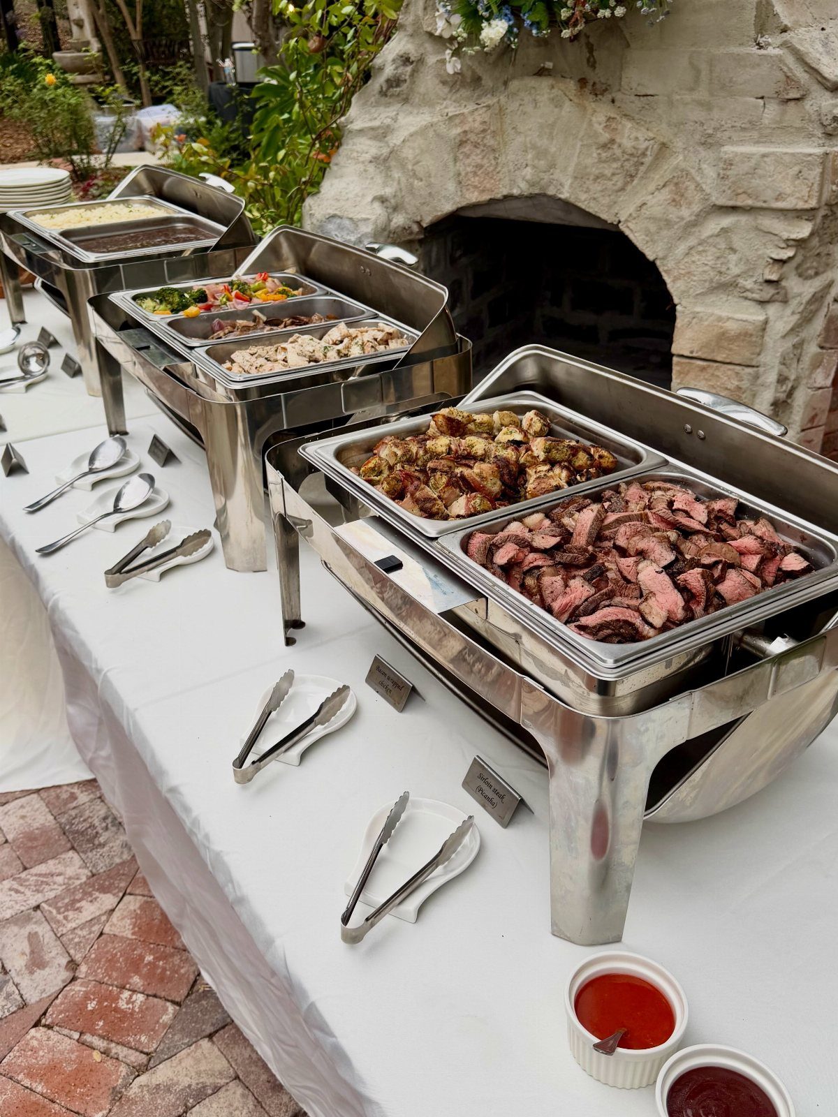 Buffet line with silver chafing dishes containing sliced meats and roasted vegetables.