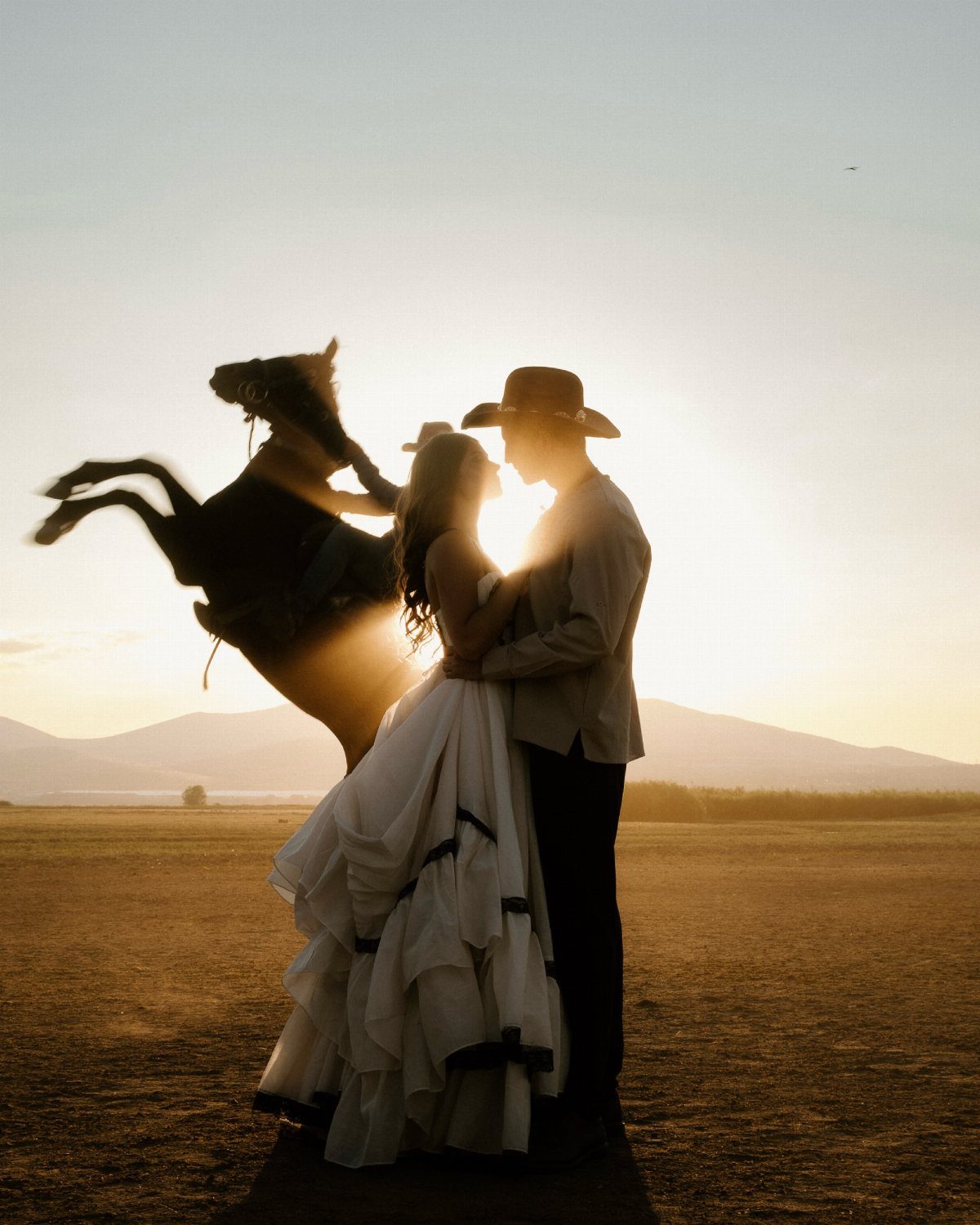 Wedding couple embracing in a field with a rearing horse silhouetted at sunset.