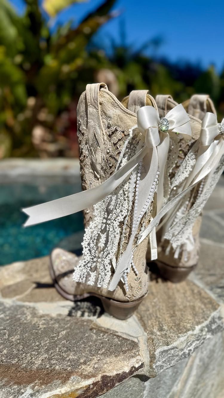 Cream cowboy boots adorned with white lace, pearls, and satin ribbons on stone.