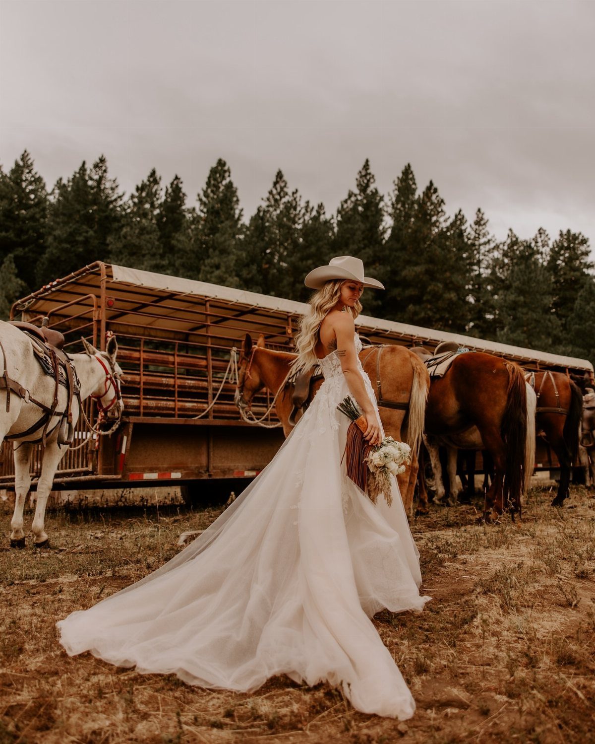 Bride in a white gown and cowboy hat standing by horses and trailer.