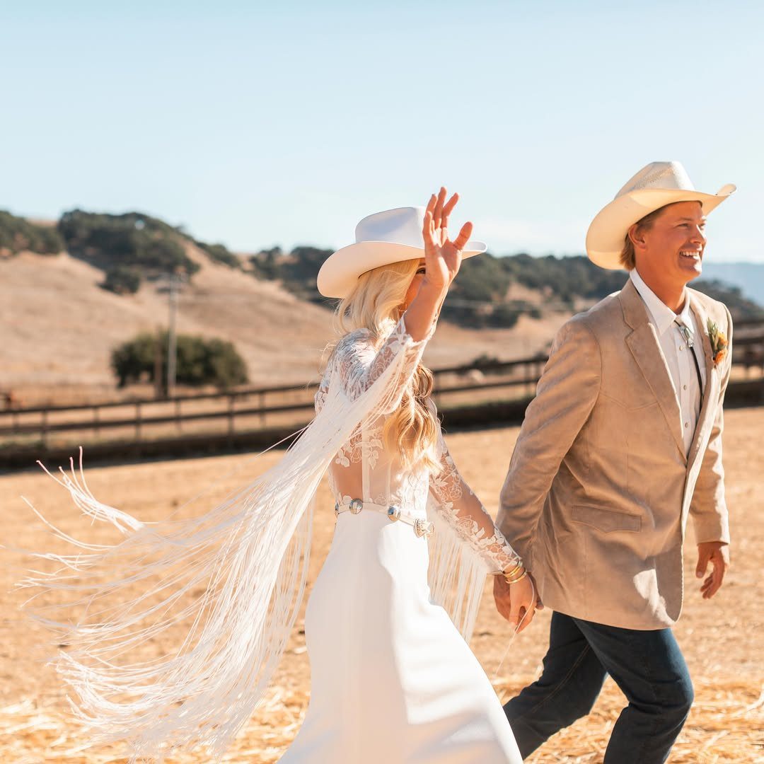 Bride in a white fringe wedding dress and cowboy hat walking with groom.