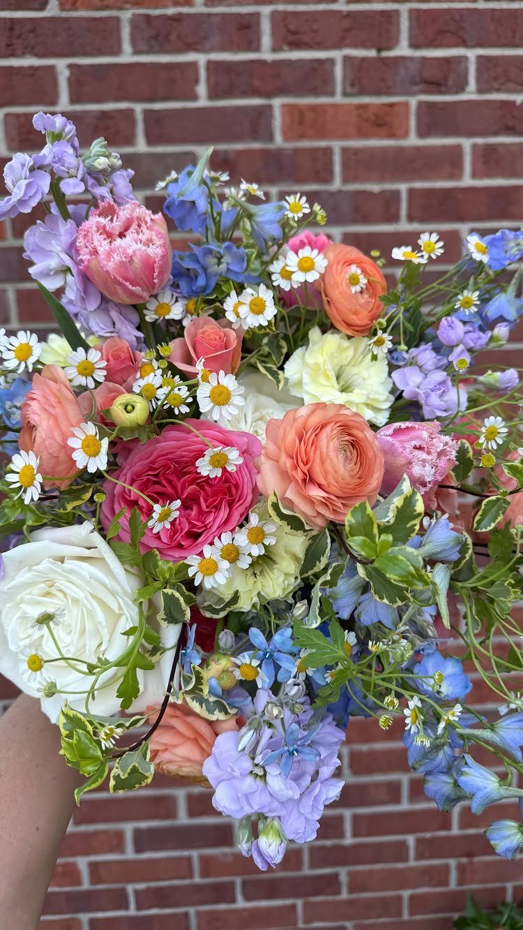 Colorful wildflower bouquet with pink, white, and blue flowers against a brick wall.