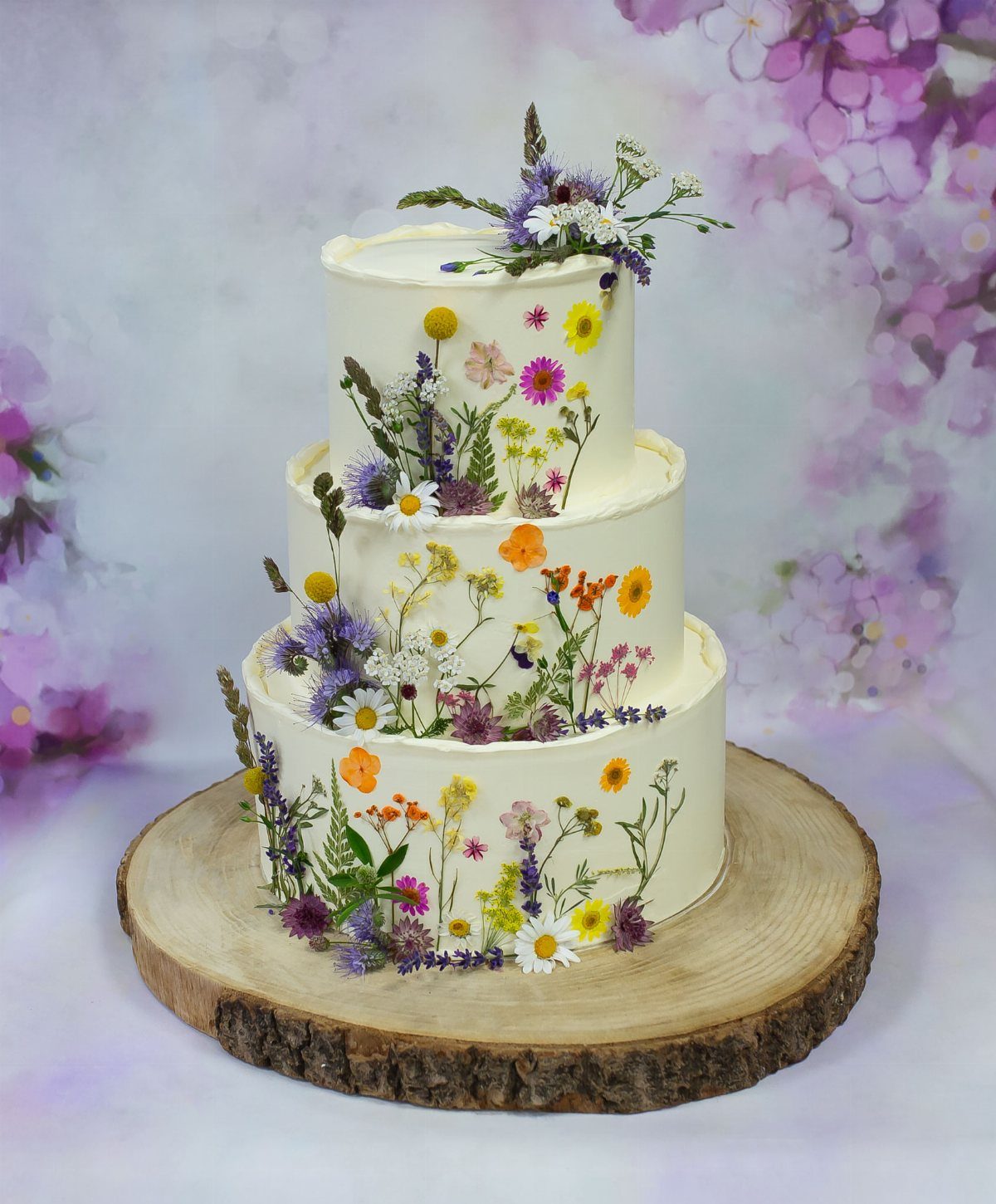 Whimsical Wildflower Wedding: 7 Decor Ideas That Feel Grown, Not Styled 8 Three-tiered white cake decorated with colorful wildflowers on a rustic wooden slice base.