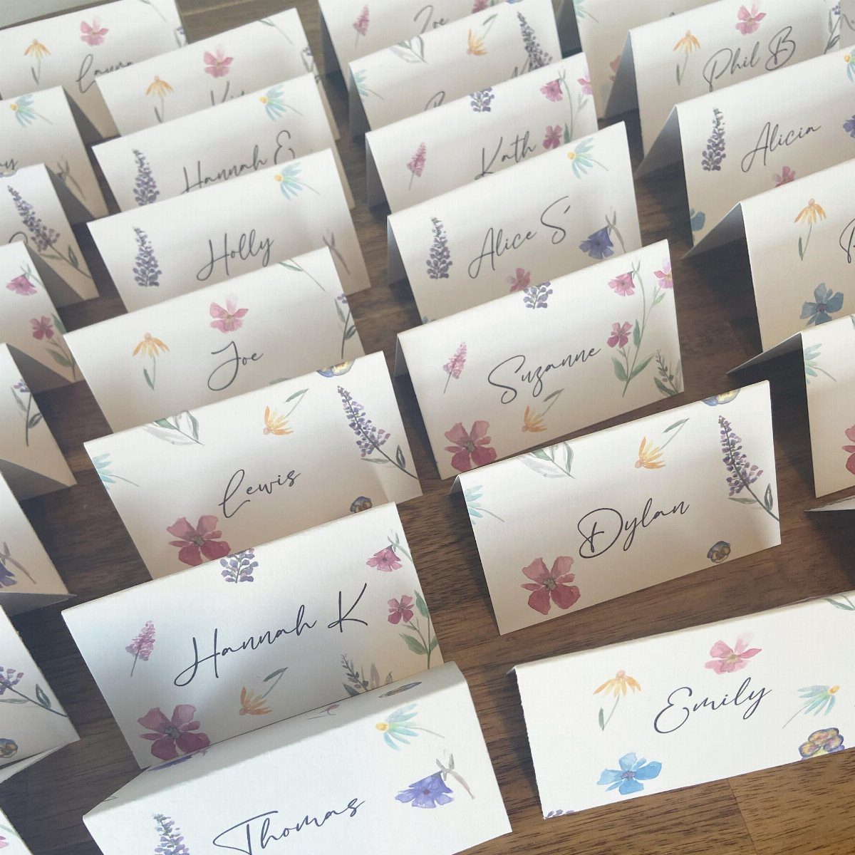 Whimsical Wildflower Wedding: 7 Decor Ideas That Feel Grown, Not Styled 6 White folded place cards with colorful wildflower illustrations and handwritten names on a wooden table.