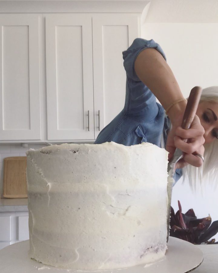 9 White Buttercream Cake Ideas That Look Bakery-Worthy at Home 4 A woman applying a thin layer of white frosting to a tall cake using a spatula.