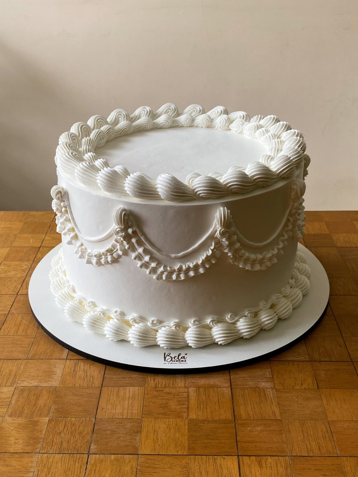 9 White Buttercream Cake Ideas That Look Bakery-Worthy at Home 6 A round white buttercream cake with elegant piped scalloped decorations on a wooden table.