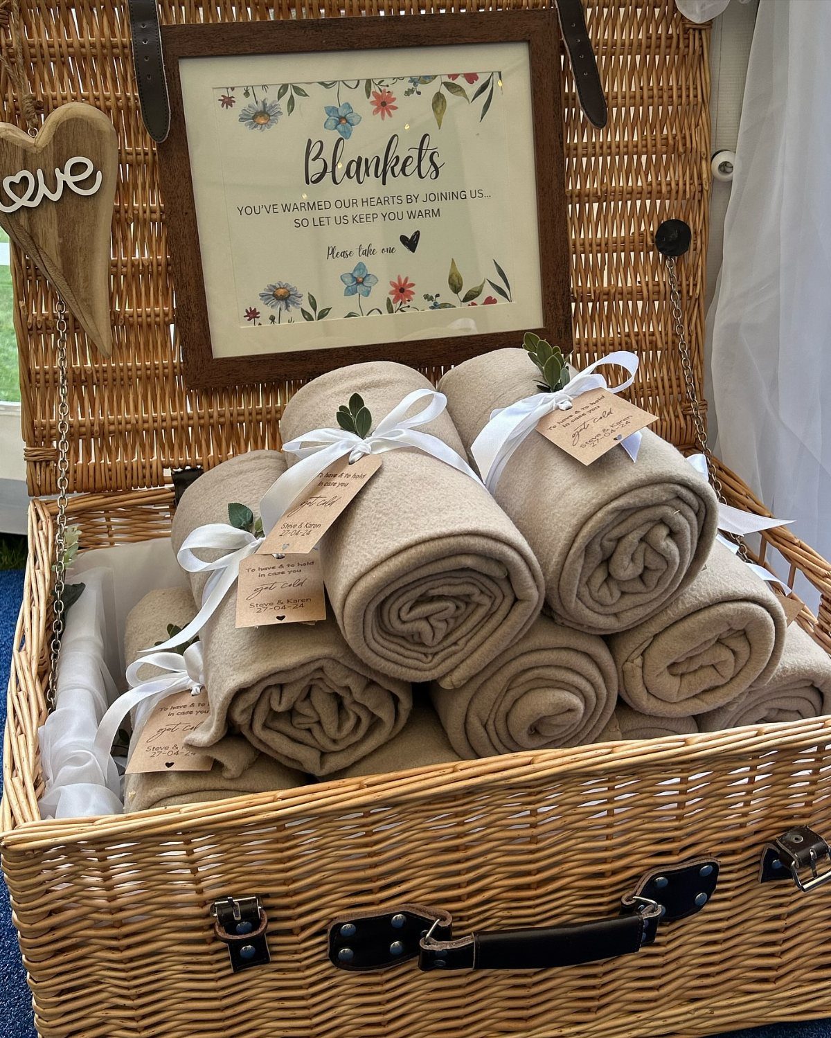 Rolled beige blankets with white ribbons inside a large wicker basket with a sign.