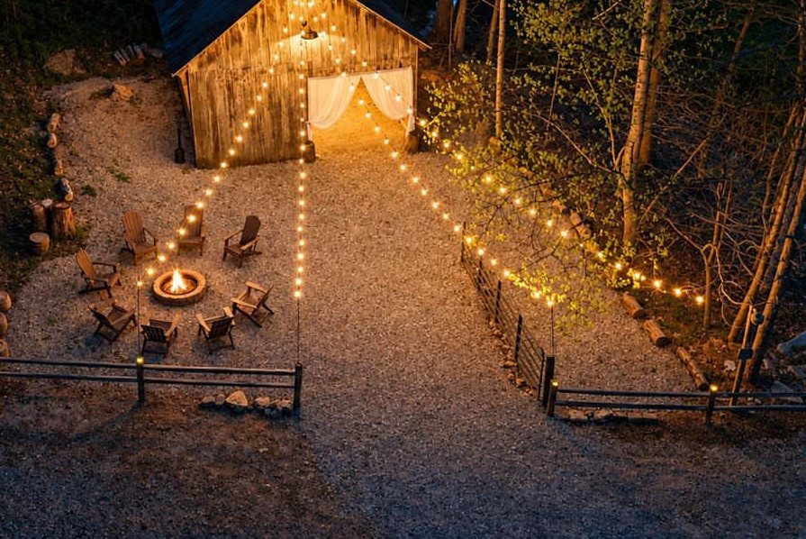 Outdoor fire pit with wooden chairs and string lights near a rustic cabin.