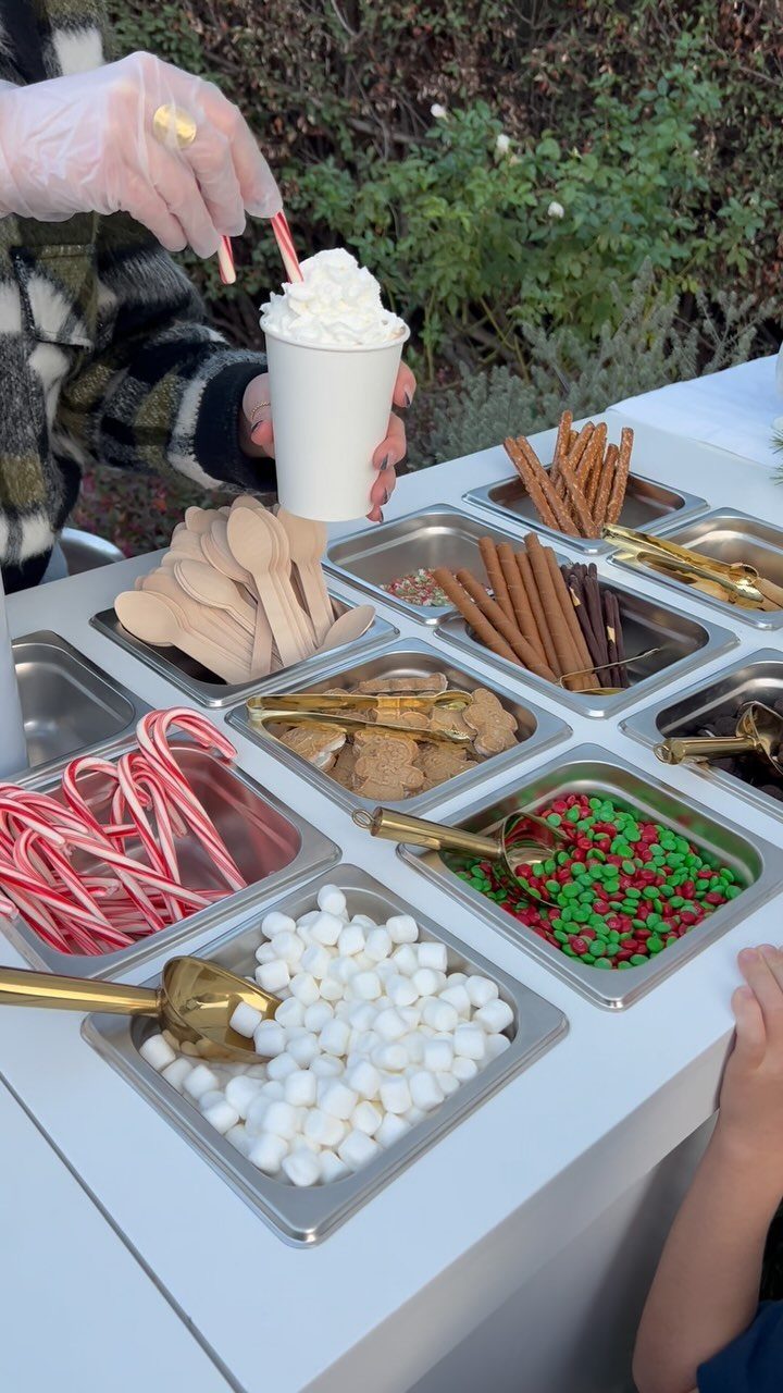 Hot cocoa bar with toppings like marshmallows, candy canes, and cinnamon sticks in trays.