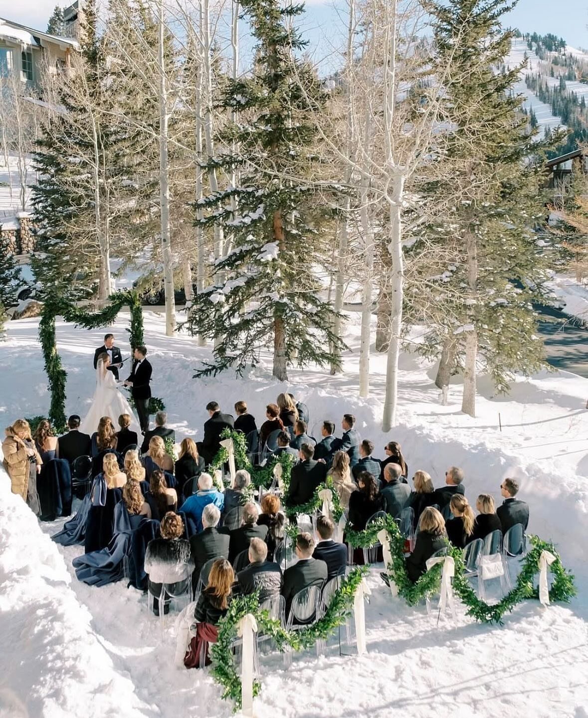 Winter wedding ceremony with guests seated in clear chairs in a snowy forest.